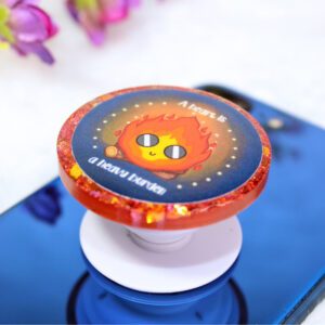 Pop socket de resina de color rojo con ilustración inspirada en Calcifer y la frase "A heart is a heavy burden"